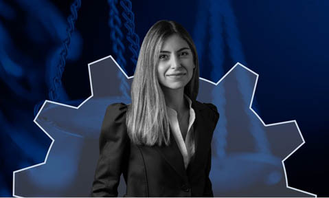 Pessoa adulta com cabelo liso e longo usando terno preto e camisa branca est em frente a um fundo azul com uma engrenagem estilizada.  Yapay zeka taraf ndan olu turulan a  klama